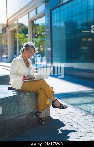 Heureuse femme d'affaires attirante d'âge moyen travaillant souriante avec un ordinateur portable devant un bâtiment moderne. Prise de vue verticale grand angle. Banque D'Images
