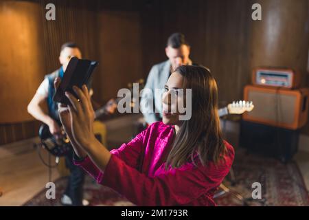 gaie chanteuse-femme en streaming d'une vidéo en direct pendant la répétition, d'autres membres du groupe dans le studio de musique de fond. Photo de haute qualité Banque D'Images