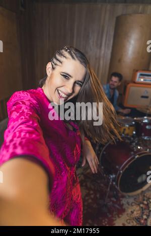 joyeux jeune chanteur en vêtements roses prenant un selfie avec un batteur, studio musical. Photo de haute qualité Banque D'Images