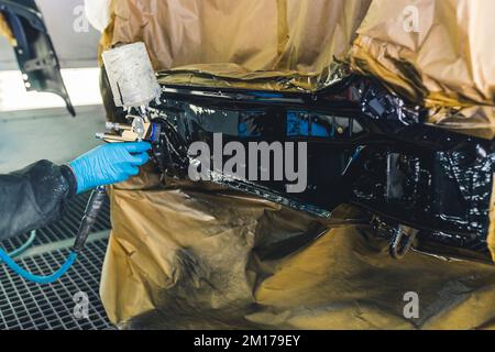 Concept de détail de voiture. À l'intérieur de la cabine de peinture. Mettre à la main des gants de protection en caoutchouc bleus tenant un pistolet à peinture et peindre une aile noire. Photo de haute qualité Banque D'Images