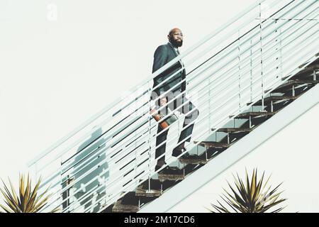 Un homme à barbe noire et chauve monte les escaliers. Le mâle africain monte un escalier métallique blanc un jour brillant, porte un costume sombre et tient un l Banque D'Images