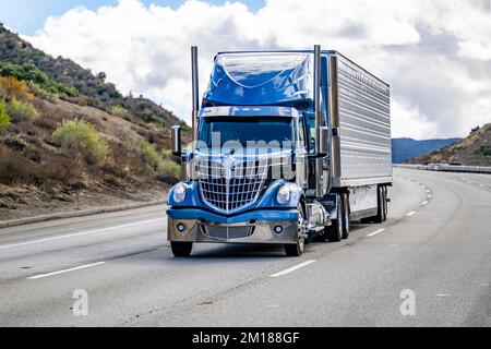 Tracteur semi-remorque bleu de grande taille de classe industrielle avec pièces chromées transportant des marchandises commerciales congelées dans un réfrigérateur semi-remorque en marche o Banque D'Images