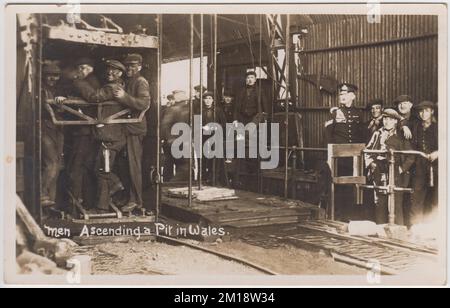 Extraction de charbon - hommes montant une fosse au pays de Galles. Photographie du début du 20th siècle d'hommes dont le visage est noirci par le charbon, entassés dans l'ascenseur de la fosse, qui se lève après la fin de leur quart de travail. Les hommes et les garçons attendent en arrière-plan pour descendre la fosse. Tout le monde porte une casquette plate, à l'exception d'un homme en uniforme Banque D'Images