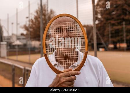 portrait d'un joueur de tennis senior vêtu de vêtements de sport posant avec une raquette sur le visage sur un court de tennis en terre battue - concept de bien-être à la retraite Banque D'Images