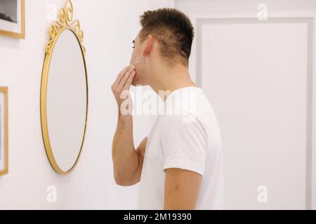 Un homme souriant applique de la crème sur le visage. Homme debout avec miroir dans un studio de beauté Banque D'Images