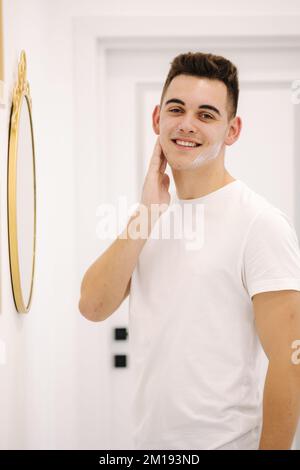 Un homme souriant applique de la crème sur le visage. Homme debout avec miroir dans un studio de beauté Banque D'Images