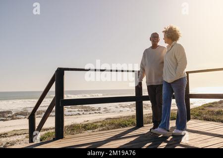 Couple d'âge mûr souriant l'un à l'autre tout en faisant une promenade le long d'un pont de pied en bois à la plage. Couple romantique de personnes âgées appréciant de dépenser la qualité t Banque D'Images
