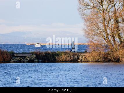 South Shore Lough Neagh, près de Lurgan, comté d'Armagh, Irlande du Nord, Royaume-Uni. 11 décembre 2022. Temps au Royaume-Uni - le froid continue avec des températures tombant à -7C dans certaines parties de l'Irlande du Nord pendant la nuit. Un peu de soleil a fait monter la température jusqu'à environ 4C à l'heure du déjeuner, mais même la brise légère est amèrement froide. Un homme marchant le long de la marina de Kinnego brise-lames lors d'une journée d'hiver ensoleillée avec de la neige dans les montagnes de Sperrin.Credit: CAZIMB/Alamy Live News. Banque D'Images