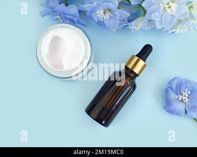 Sérum dans un flacon en verre brun sans marque avec pipette, crème hydratante dans un pot en verre ouvert et fleur de delphinium bleue sur fond bleu. Bouteille cosmétique Banque D'Images