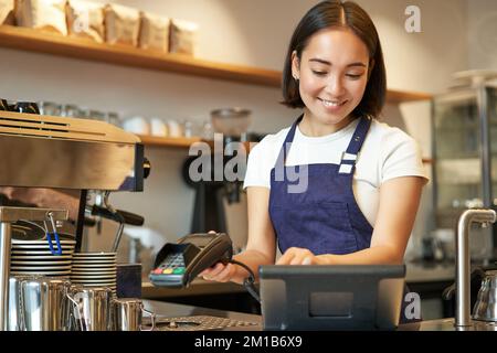 Une femme de barista asiatique souriante traite la commande, entre la commande dans le terminal de point de vente et travaille au comptoir dans le café-restaurant Banque D'Images