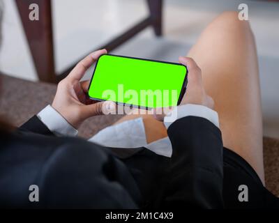 Écran vert sur téléphone mobile de style horizontal dans les mains de femme d'affaires qui portent un costume et un short formels, assis détendu avec des jambes croisées sur le canapé en co Banque D'Images