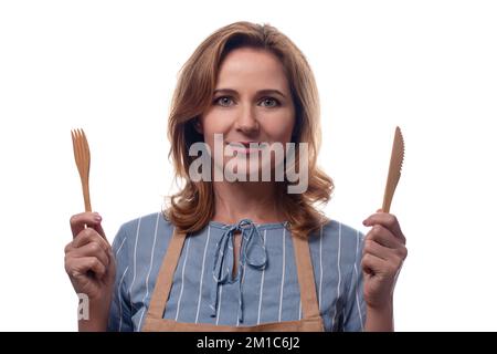femme caucasienne en tablier tenant une fourchette et un couteau en bois, isolée sur fond blanc. Ustensiles de cuisine jetables écologiques. Kit pique-nique. Pas de pla Banque D'Images