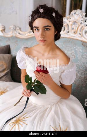 Portrait jeune belle mariée attrayante avec rose rouge dans sa main. Robe blanche vintage et bijoux de mariée. Assis sur le canapé Banque D'Images