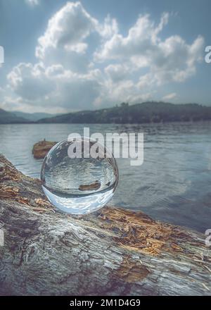 Un cliché vertical d'une boule de cristal sur une bûche d'arbre tombée, reflétant un lac tranquille par une journée nuageux Banque D'Images