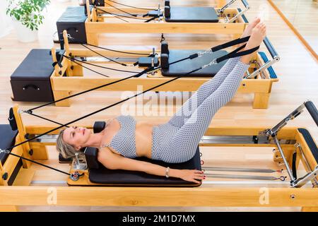 De haut pieds nus femme dans les vêtements de sport levant les jambes et faisant cent exercice sur le lit reformeur pendant l'entraînement dans le studio pilates léger Banque D'Images