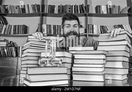 Homme sur le visage heureux entre des piles de livres, tout en étudiant dans la bibliothèque, les étagères sur le fond. Un professeur ou un étudiant avec une barbe est assis à table avec des livres Banque D'Images