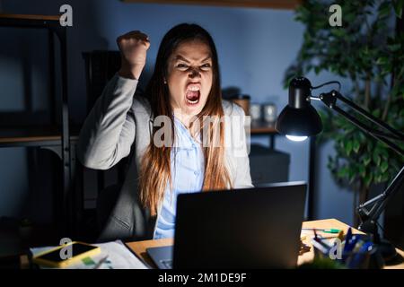 Jeune femme brune travaillant au bureau la nuit en colère et folle levant poing frustré et furieux tout en criant avec colère. Rage et agressif c Banque D'Images