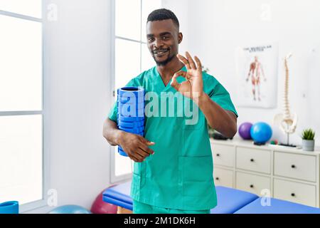 Jeune homme kinésithérapeute africain tenant le rouleau de mousse à la clinique faisant signe ok avec les doigts, souriant sympathique gestant excellent symbole Banque D'Images