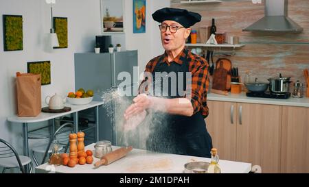 Baker portant un uniforme jette la farine debout près de la table. Cuisinier professionnel avec des crottes de bonete et de la mouche de poudre, chef à la retraite parsemant des ingrédients rew sur la pâte préparant le pain traditionnel. Banque D'Images