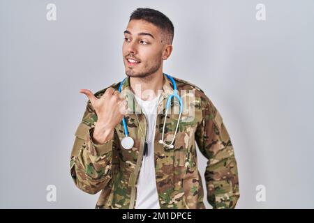 Jeune médecin hispanique portant l'uniforme militaire de camouflage pointant le pouce vers le côté souriant heureux avec la bouche ouverte Banque D'Images