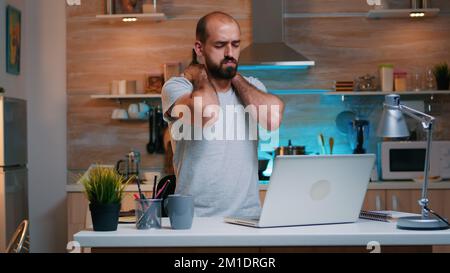 Un homme épuisé prend une pause de travail à distance et stretching son cou assis devant l'ordinateur portable. Employé occupé utilisant le réseau de technologie moderne sans fil travail heures supplémentaires lecture écriture. Banque D'Images