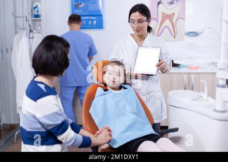 Le spécialiste de la dentistirie parle de l'hygine buccale avec les jeunes parents et les enfants. Stomatolog expliquant la prévention des dents à la mère et à l'enfant tenant un Tablet pc avec l'espace de copie disponible. Banque D'Images