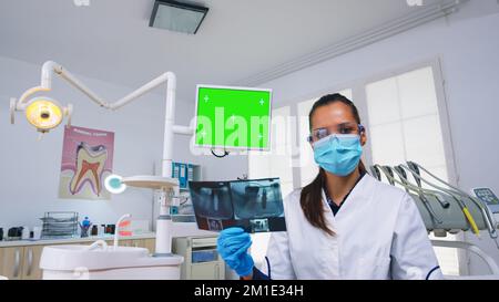 Dentiste médecin montrant le moniteur de l'unité avec écran maquette, tenant la radiographie des dents et examinant l'infection des dents. Spécialiste en Stomatology avec masque facial pointant vers l'affichage chromique de l'espace de copie de l'écran vert Banque D'Images