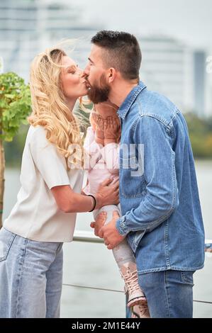 Homme barbu tenant une petite fille et embrassant une femme blonde dans la rue. Enfant mignon couvrant les yeux avec les mains tandis que les parents partagent le moment tendre à l'extérieur. Concept de famille et d'amour. Banque D'Images