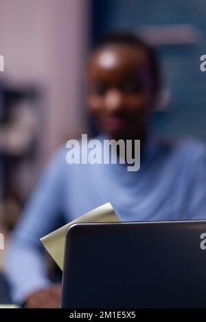 Des notes adhésives sur ordinateur portable avec une femme d'affaires africaine rouée en arrière-plan. Travailleur indépendant noir respectant la date limite pour étudier tard dans la nuit. Banque D'Images