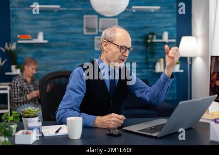 Un homme senior enthousiaste qui célèbre les bonnes nouvelles tout en travaillant sur un ordinateur portable à domicile. Homme âgé entrepreneur dans le lieu de travail à la maison utilisant un ordinateur portable assis au bureau tandis que la femme lit un livre assis sur le canapé. Banque D'Images