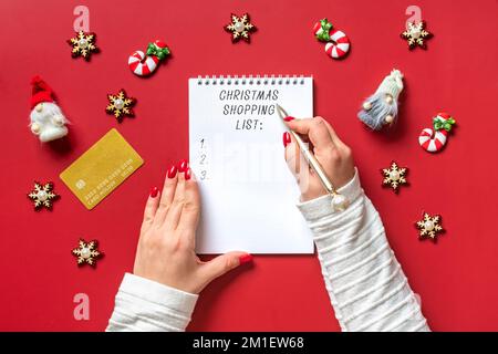 La main féminine tient un stylo et écrit la liste d'achats de Noël, idées de cadeau sur le bloc-notes blanc sur fond rouge vue du dessus plat Lay carte de vacances femme faisant Noël liste de cadeaux, planification, concept d'achat. Banque D'Images