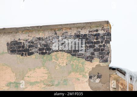 Un mur de sorbets avec des briques brûlées et des glaçons sur le bord. Un vieux bâtiment avec du plâtre abîmé contre un ciel gris. De grandes cascades de glaces pendent d'affilée. Jour d'hiver nuageux, lumière douce. Banque D'Images