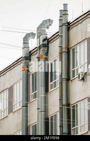 Métal, longs tuyaux de ventilation d'échappement sur le mur de la maison avec des fenêtres. Le toit de la maison dans la neige contre le ciel gris. Nuageux, froid jour d'hiver, lumière douce. Banque D'Images