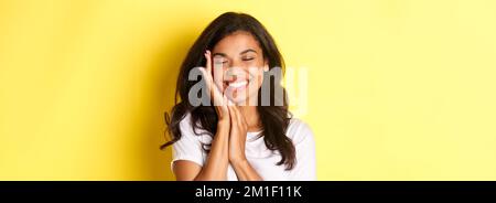 Gros plan d'une femme afro-américaine rêveuse, au visage doux et souriante, debout sur fond jaune Banque D'Images