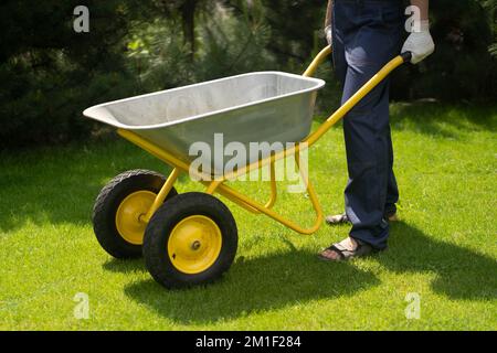 Un jeune homme avec les mains en gants porte une voiturette de jardin en métal Banque D'Images