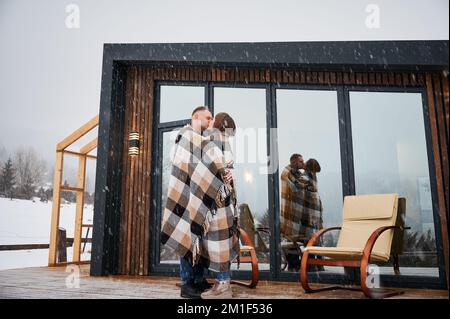 Homme et femme enveloppés dans une couverture embrassant à l'extérieur sous la neige d'hiver. Couple heureux partageant un moment romantique tout en se tenant près des chaises à l'extérieur de la maison de vacances scandinave avec des fenêtres panoramiques. Banque D'Images