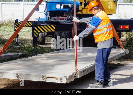 Le Slinger pose une dalle de béton sur le chantier de construction le jour de l'été. Le travailleur dans la veste de protection et le casque de construction supervise la pose de la base sur le chantier. Banque D'Images