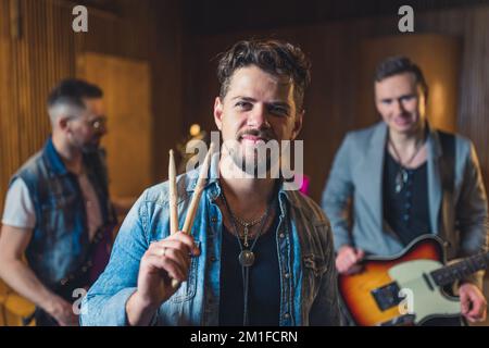 jeune batteur à poil dur tenant des tambours bâtons deux guitaristes en arrière-plan, studio d'enregistrement. Photo de haute qualité Banque D'Images