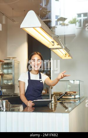 Une jeune femme de barista asiatique heureuse travaille dans un café, fait des vagues au client, traite les commandes derrière le comptoir dans un café, porte un tablier bleu Banque D'Images