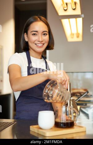 Cliché vertical du barista coréen souriant, en versant du café filtre, en préparant une préparation en lot pour le client dans le café, en versant la méthode Banque D'Images