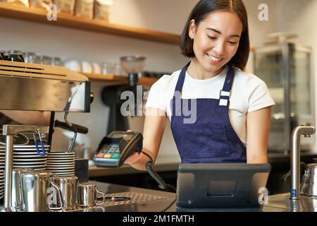 Une femme de barista asiatique souriante traite la commande, entre la commande dans le terminal de point de vente et travaille au comptoir dans le café-restaurant Banque D'Images