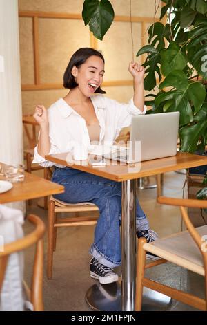 Une jeune fille asiatique enthousiaste qui célèbre, regarde un ordinateur portable et triomphant, fait des pompes de poing avec le visage excité, se trouve dans un café Banque D'Images