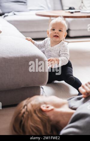 Des moments heureux en famille. Mère couchée confortablement sur le tapis des enfants jouant avec son bébé garçon en regardant et en défendant ses premiers pas. Émotions humaines positives, sentiments, joie Banque D'Images