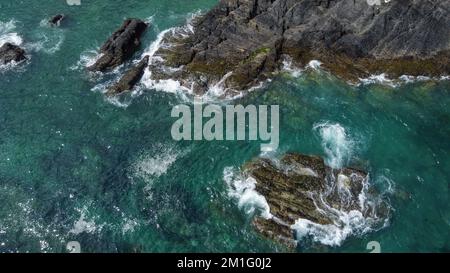 Rochers couverts de mousse noire au milieu des vagues turquoise de la mer celtique. Mousse de mer blanche sur les vagues. Eaux de l'océan Atlantique. Point de vue du drone. Banque D'Images