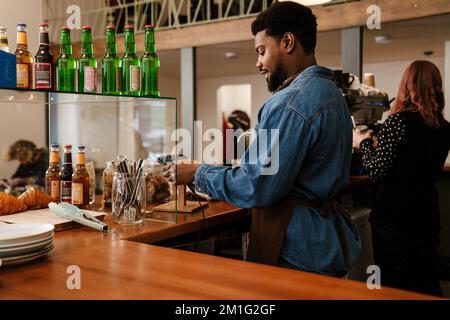 Homme à barbe noire portant un tablier travaillant à la caisse dans un café à l'intérieur Banque D'Images