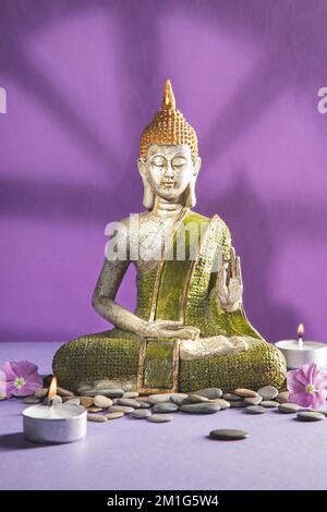 Statue de bouddha verte et dorée en méditation avec ombres sur fond violet clair. Format vertical. Banque D'Images