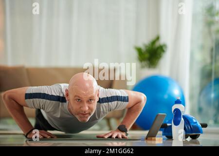 Homme d'âge moyen qui fait des push-ups lors de l'entraînement à la maison avec un instructeur de fitness en ligne Banque D'Images