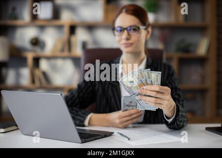 Concentrez-vous sur la main, tenant un paquet d'argent, de la jeune femme d'affaires caucasienne de cheveux rouges utilisant un ordinateur portable comptant les dollars américains dans le nouveau bureau moderne. Banque D'Images
