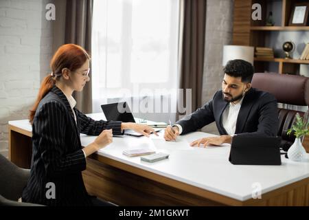 Homme d'affaires confiant en costume noir donne un salaire à une jeune femme d'affaires de cheveux rouges. Un prêt en espèces sur salaire. Chef d'entreprise payant de l'argent comptant de nouveaux euros et des dollars aux employés de bureau. Banque D'Images