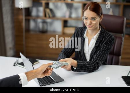 Vue écourtée d'un homme d'affaires non reconnu qui a donné des lots de billets de cent dollars à une jeune femme pdg en cheveux rouges. Femme d'affaires recevant de l'argent de l'homme d'affaires. Vénalité, pot-de-vin, concept de corruption. Banque D'Images